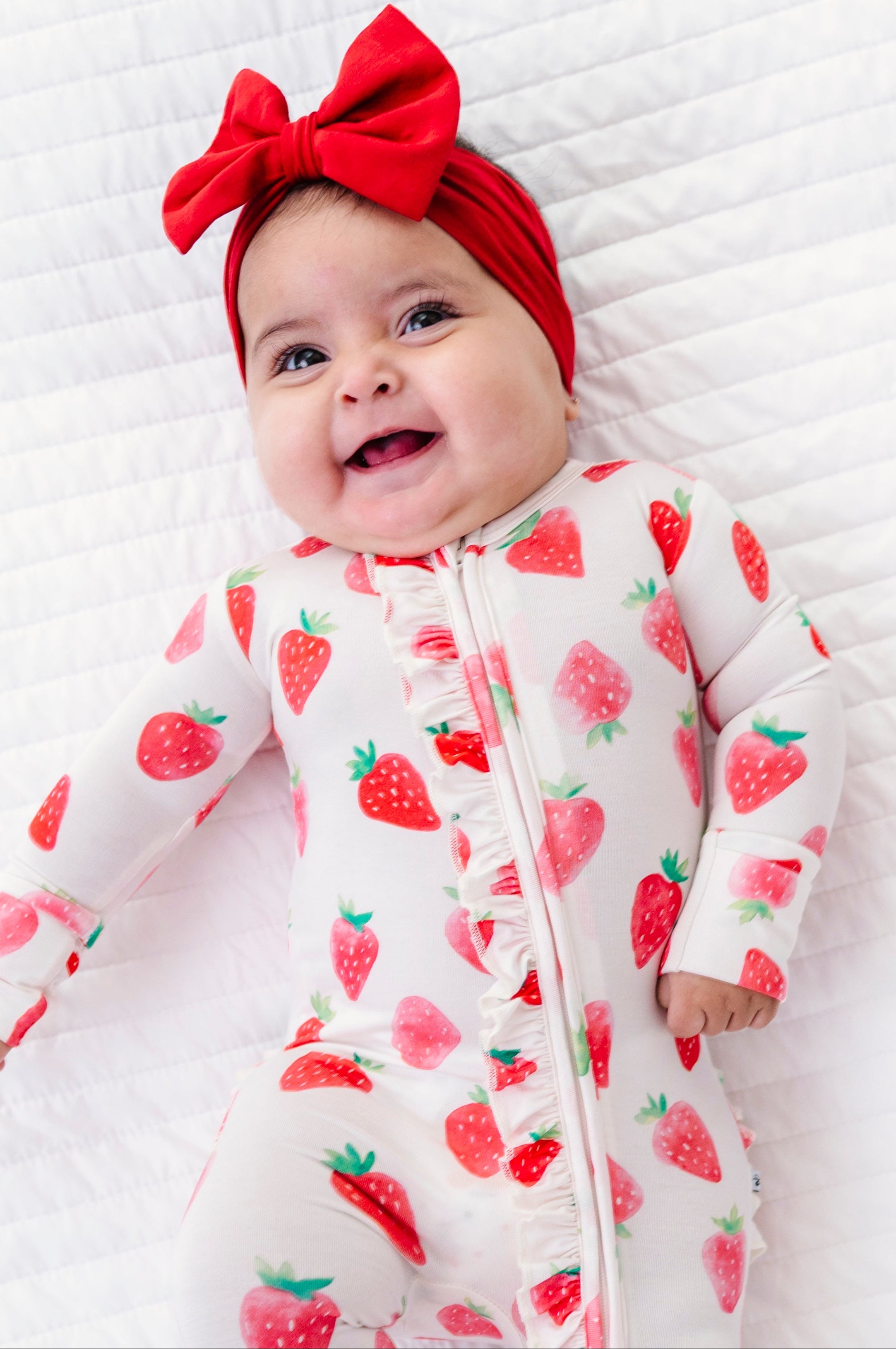 Baby wearing a strawberry-patterned onesie and red headband on a white quilted surface